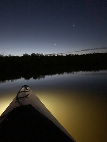 Bow of a kayak with a light on the front on a river at night.