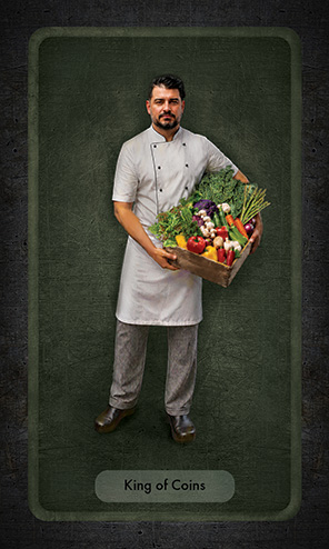 The King of Coins shows a professional chef in a white coat, white half apron, black gingham checked pants and black clogs holding a large wooden box of many different colorful vegetables.