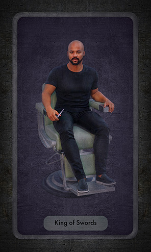 The King of Swords is a strong black man dressed in a black t-shirt and black jeans sitting in a barber's chair .holding a pair of scissors.