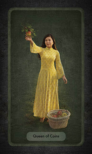 The Queen of Coins wears a long, Asian-style tunic in yellow, embroidered with the Chinese symbol for longevity. She picks an apple from a low-hanging branch to place in the nearly-full basket beside her feet.