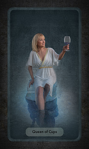 The Queen of Cups shows a blond woman seated on a rock, her feet and lower leg submersed in the pool that surrounds her. Her left hand holds a glass of red wine raised in a toast.